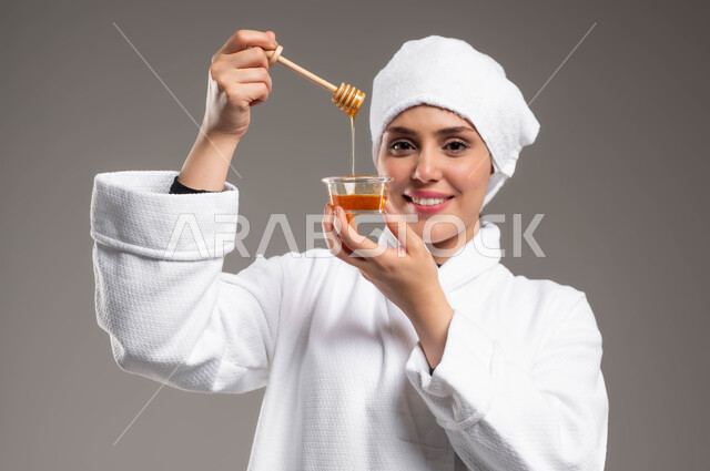 النظر للكاميرا بايماءات السرور والسعادة، اتباع حمية غذائية، استخدام عسل النحل والمنتجات الطبيعية للعناية بالبشرة، منتج وطني محلي سعودي، بورتريه مقرب لامرأة عربية خليجية سعودية مبتسمة ترتدي روب استحمام ومنشفة بيضاء تحمل بيدها ملعقة خشبية مغمورة بالعسل، خلفية رمادية
