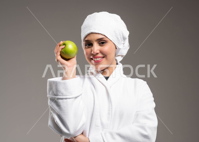 فطور وسناك صحي مفيد، حمية غذائية صحية، فيتامينات مغذية للجسم، الفاكهة الطازجة المفيدة للصحة، اتباع نظام غذائي صحي، بورتريه مقرب لمرأة عربية خليجية سعودية ترتدي روب استحمام ومنشفة بيضاء تحمل بيدها تفاحة خضراء طازجة، خلفية رمادية