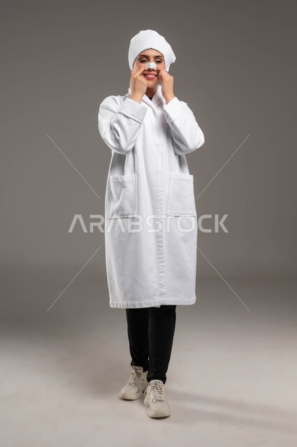 التخلص من الرؤوس السوداء و الجلد الميت، استخدام ماسكات تنظيف المسام ومكونات تجميلية، مفهوم العناية والاهتمام بنضارة وتحسين البشرة، بورتريه لامرأة عربية خليجية سعودية ترتدي روب استحمام أبيض و منشفة بيضاء تضع لصقات الأنف،صورة لكامل الجسم، خلفية رمادية