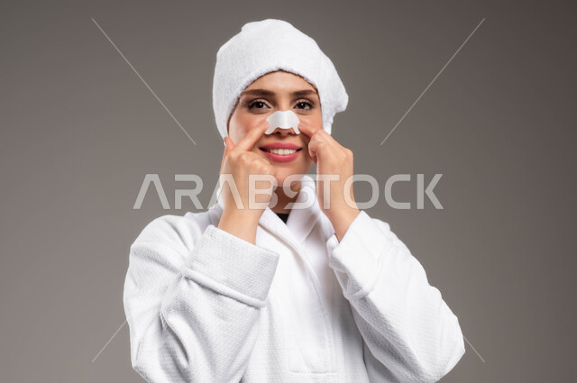 التخلص من الرؤوس السوداء و الجلد الميت، استخدام ماسكات تنظيف المسام ومكونات تجميلية، مفهوم العناية والاهتمام بنضارة وتحسين البشرة، بورتريه مقرب لامرأة عربية خليجية سعودية ترتدي روب استحمام أبيض و منشفة بيضاء تضع لصقات الأنف، خلفية ملونة