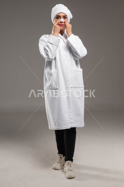 استخدام ماسكات تنظيف المسام ومكونات تجميلية، مفهوم العناية والاهتمام بنضارة وتحسين البشرة، التخلص من الرؤوس السوداء والجلد الميت، بورتريه لامرأة عربية خليجية سعودية ترتدي روب استحمام أبيض و منشفة بيضاء تضع لصقات الأنف، صورة لكامل الجسم، خلفية ملونة