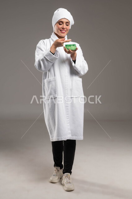 الاستعداد للاهتمام بنظافة و نضارة البشرة، تنظيف و تقشير الوجه، استخدام مكونات طبيعية للعناية بالمظهر الخارجي،  بورتريه لامرأة عربية خليجية سعودية مبتسمة ترتدي روب استحمام أبيض ومنشفة بيضاء تقوم بوضع جل مرطب، صورة الجسم بالكامل، خلفية رمادية