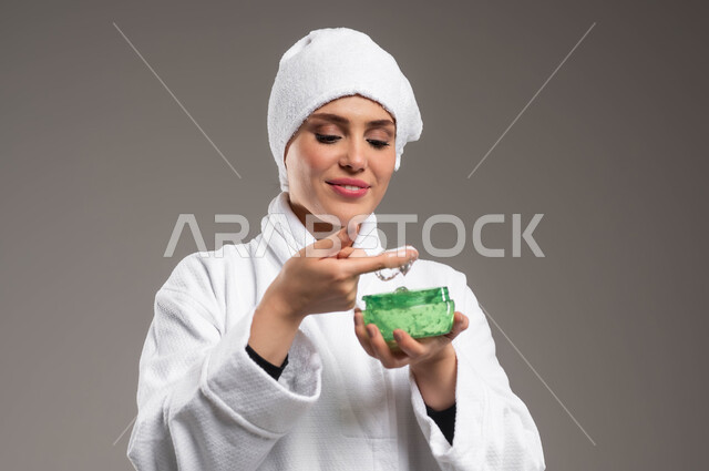 استخدام مكونات تجميلية للعناية بالمظهر الخارجي، الاستعداد للاهتمام بنظافة و نضارة البشرة، تنظيف و تقشير الوجه، بورتريه مقرب لامرأة عربية خليجية سعودية مبتسمة ترتدي روب استحمام أبيض ومنشفة بيضاء تقوم بوضع جل مرطب، خلفية رمادية