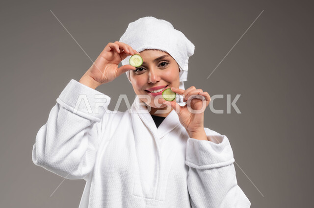 النظر إلى الكاميرا بإيماءات السرور و السعادة، الاستعداد للاهتمام بنظافة ونضارة البشرة، استخدام المكونات الطبيعية للعناية بالمظهر الخارجي، تنظيف وتقشير الوجه، بورتريه مقرب لامرأة عربية خليجية سعودية مبتسمة ترتدي روب استحمام أبيض ومنشفة بيضاء تحمل بيديها قطع من الخيار، خلفية رمادية