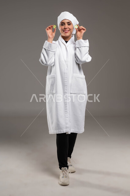 استخدام مكونات طبيعية للعناية بالمظهر الخارجي، الاستعداد للاهتمام بنظافة ونضارة البشرة، تنظيف وتقشير الوجه، بورتريه لامرأة عربية خليجية سعودية مبتسمة ترتدي روب استحمام أبيض ومنشفة بيضاء تحمل بيديها قطع من الخيار، صورة الجسم بالكامل، خلفية رمادية