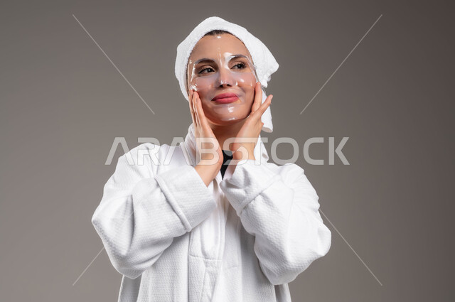 استخدام مكونات و مستحضرات تجميلية، تنظيف وتقشير الوجه، بورتريه مقرب لامرأة عربية خليجية سعودية ترتدي روب استحمام و منشفة بيضاء تقوم بوضع قناع النضارة على وجهها، روتين العناية بالبشرة، مفهوم الاهتمام بالمظهر الخارجي عند النساء، خلفية رمادية