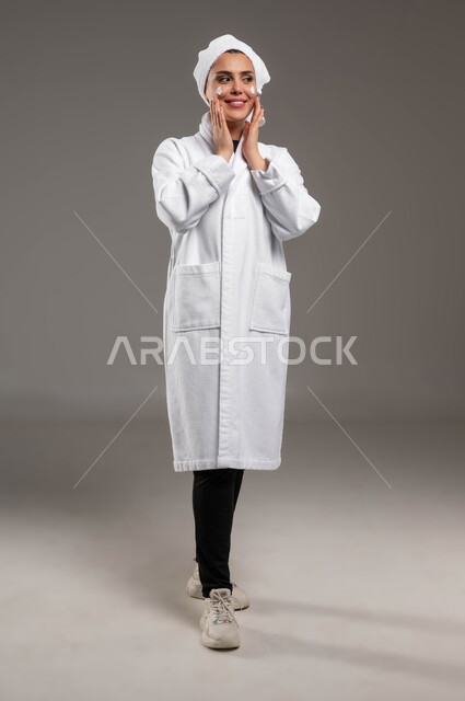مفهوم العناية بنضارة وتحسين البشرة، الاهتمام بالمظهر الخارجي، بورتريه لامرأة عربية خليجية سعودية ترتدي روب استحمام أبيض و منشفة بيضاء تضع كريم الترطيب بإيماءات السرور، استخدام كريمات ومكونات تجميلية، صورة الجسم بالكامل، خلفية رمادية