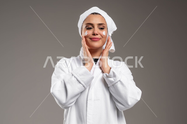 تدليك و ترطيب الوجه، الاهتمام بالمظهر الخارجي، مفهوم العناية بنضارة وتحسين البشرة، بورتريه مقرب لامرأة عربية خليجية سعودية مبتسمة ترتدي روب استحمام أبيض و منشفة بيضاء تضع كريم مرطب، استخدام كريمات و مكونات تجميلية، خلفية رمادية
