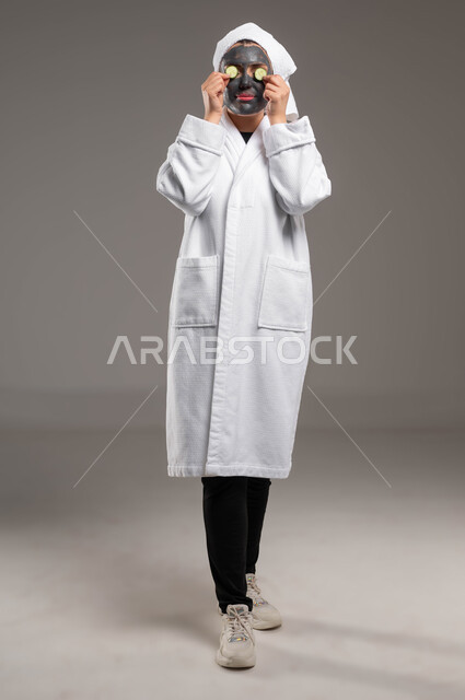 مفهوم العناية والاهتمام بالمظهر الخارجي عند النساء، استخدام مكونات و مستحضرات تجميلية، خطوات تنظيف وتقشير البشرة، بورتريه لامرأة عربية خليجية سعودية ترتدي منشفة وروب الاستحمام الابيض تقوم بوضع قناع الطين وقطع الخيارعلى وجهها، صورة الجسم بالكامل، خلفية رمادية
