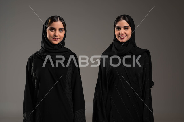 إيماءات الفرح و السرور، الوقوف باستقامة والنظرإلى الكاميرا، بورتريه مقرب لفتاتين توءم عربيتين سعوديتين خليجيتين مبتسمتين  محجبتين ترتديين عباءة سوداء، مشاركة اللحظات السعيدة، التشارك واللعب مع الأخوات، قضاء أوقات ممتعة ومسلية، خلفية رمادية