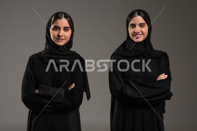 الوقوف باستقامة والنظر إلى الكاميرا بايدي متقاطعة، بورتريه مقرب لفتاتين توءم عربيتين سعوديتين خليجيتين محجبتين تنظران بإيماءات السرور والسعادة، التعاطف و المودة بين الشقيقات، مفهوم العلاقة بين الأخوات، قضاء الوقت مع العائلة، تشارك اللحظات الممتعة، خلفية رمادية