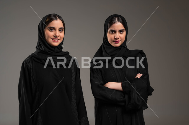 النظر إلى الكاميرا بإيماءات وجه و يدين مختلفة، بورتريه مقرب لفتاتين توءم عربيتين سعوديتين خليجيتين محجبتين تنظر إحداهما بغضب و الاخرى بابتسامة و سعادة، مفهوم الصداقة و الأخوة، العلاقة بين الأخوات، قضاء الوقت مع العائلة، خلفية رمادية