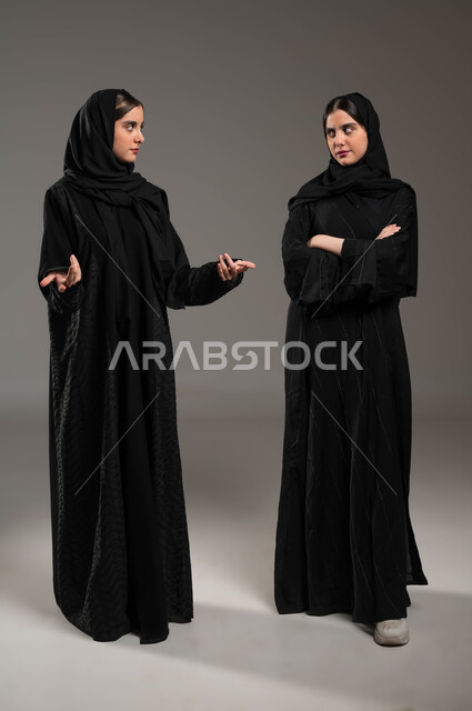 Looks of hatred and anger, a portrait of two veiled Saudi Gulf Arab twin girls, one of whom looks angrily and maliciously and the other makes questioning and questioning movements, the concept of body language, the concept of the relationship between sisters, a full-length body image, gray background.