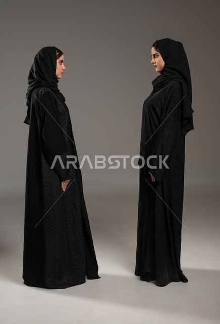 تعابير المواجهة بين الاخوات, النظر الى بعض بثقة, ايماءات التحدي والاصرار, مفهوم لغة الجسد والحركات, بورتريه من الجانب لبنتان عربيتان سعوديتان خليجيتان ترتديان العباءة والحجاب, صورة الجسم بالطول الكامل, خلفية رمادية