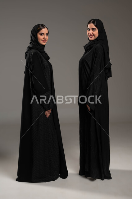 ايماءات التحدي والاصرار, تعابير المواجهة بين الاخوات, النظر الى الكاميرا بثقة والابتسامة, مفهوم لغة الجسد والحركات, بورتريه من الجانب لبنتان عربيتان سعوديتان خليجيتان ترتديان العباءة والحجاب, صورة الجسم بالطول الكامل, خلفية رمادية