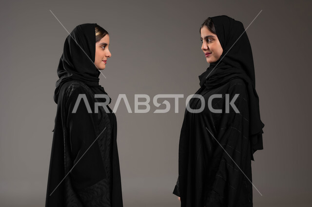النظر الى بعض بثقة, تعابير المواجهة بين الاخوات, مفهوم لغة الجسد والحركات, بورتريه مقرب من الجانب لبنتان عربيتان سعوديتان خليجيتان ترتديان العباءة والحجاب, ايماءات التحدي والاصرار, خلفية رمادية