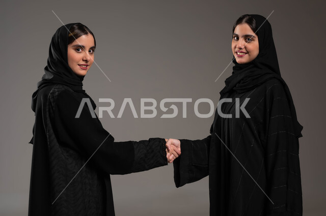 الوقوف بمحبة والنظر الى الكاميرا, السلام والمصافحة باليد, بورتريه مقرب لبنتان عريتان سعوديتان خليجيتان ترتديان الحجاب والعباءة, ايماءات الرضى والفرح, خلفية رمادية