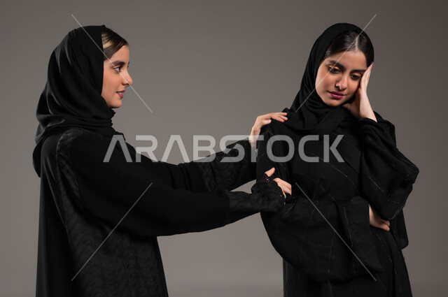 تخفيف الالم والمواساة, الوقوف بجانب بعضهم البعض, التعاطف والمودة بين الشقيقات, بورتريه مقرب لبنتان توءم عربيتان سعوديتان خليجيتان ترتديان الحجاب والعباءة احداهما تضع يدها على كتف اختها والاخرى في حالة حزن, خلفية رمادية