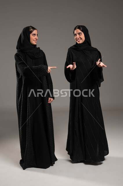 Spontaneous movements indicating something, gestures of joy and happiness, looking at each other, portrait of two Saudi Gulf Arab girls wearing the hijab and abaya, one of them pointing with her index finger at her sister and the other in confusion, full-length body portrait, gray background