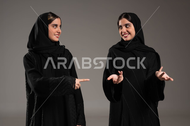 ايماءات الفرح والسعادة, حركات عفوية تدل على امر ما, النظر الى بعضهم البعض, بورتريه مقرب لبنتان عربيتان سعوديتان خليجيتان ترتديان الحجاب والعباءة احراهما تشير بأصبع السبابة على اختها والاخرى في حيرة, خلفية رمادية