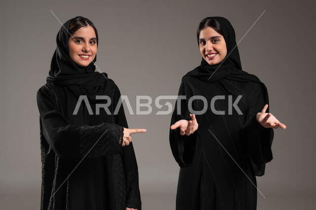 حركات عفوية تدل على امر ما, الوقوف باستقامة والنظر الى الكاميرا, بورتريه مقرب لبنتان توءم عربيتان سعوديتان خليجيتان ترتديان الحجاب والعباءة احرهما تشير بأصبع السبابة على اختها والاخرى في حيرة, خلفية ملونة