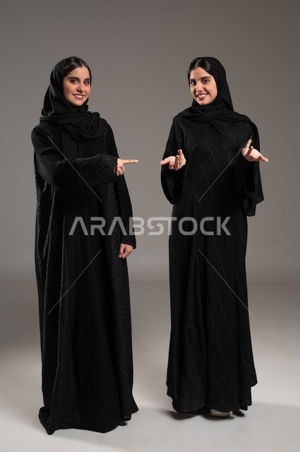 الوقوف باستقامة والنظر الى الكاميرا, حركات عفوية تدل على امر ما, بورتريه لبنتان توءم عربيتان سعوديتان خليجيتان ترتديان الحجاب والعباءة احرهما تشير بأصبع السبابة على اختها والاخرى في حيرة, صورة الجسم بالطول الكامل, خلفية ملونة