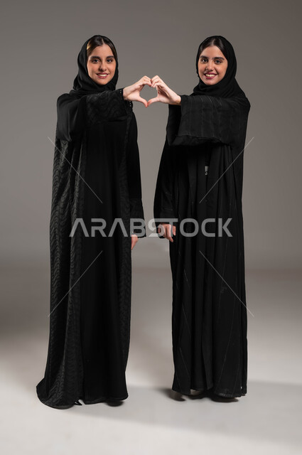 ايماءات تدل على الشعور بالحب، المحبة والترابط والألفة, العلاقة الايجابية الاخوية والحب والمودة، إشارات يدين على شكل قلب, بورتريه لتوأم بنات عربيات خليجيات سعوديات ترتديان العباءة والحجاب, صورة الجسم بالطول الكامل, خلفية رمادية