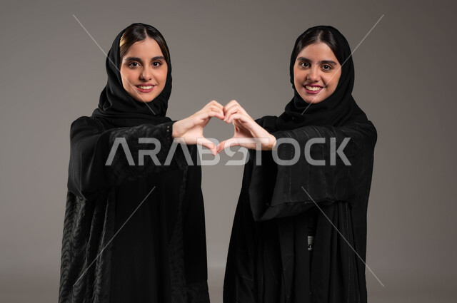 المحبة والترابط والألفة, العلاقة الايجابية الاخوية والحب والمودة، إشارات يدين على شكل قلب, ايماءات تدل على الشعور بالحب، بورتريه مقرب لتوأم بنات عربيات خليجيات سعوديات ترتديان العباءة والحجاب, خلفية رمادية