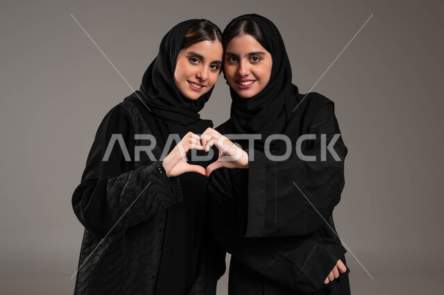 العلاقة الايجابية الاخوية والحب والمودة، إشارات يدين على شكل قلب, ايماءات وجه ويدين تدل على الشعور بالحب، بورتريه مقرب لتوأم بنات عربيات خليجيات سعوديات ترتديان العباءة والحجاب, خلفية رمادية