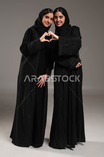 إشارات يدين على شكل قلب حب بايماءات وجه ويدين تدل على الشعور بالحب، بورتريه لتوأم بنات عربيات خليجيات سعوديات ترتديان العباءة والحجاب، العلاقة الايجابية الاخوية والحب والمودة، صورة الجسم بالطول الكامل، خلفية رمادية