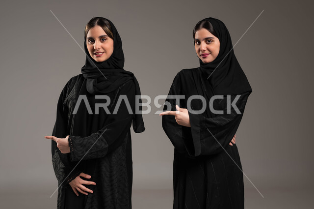 مفهوم لغة الجسد والحركات والاشارات, الاشارة الى شيء ما بأصبع السبابة, تعابير تدل على المكان, بورتريه مقرب لبنتان توءم عربيتان سعوديتان خليجيتان ترتديان الحجاب والعباءة, خلفية ملونة