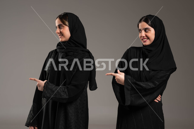الإشارة إلى شيء ما بأصبع السبابة، تحديد موقع معين بإيماءات السرور والبشاشة، مفهوم لغة الجسد والحركات والإشارات، بورتريه مقرب لبنتان توءم عربيتان خليجيتان سعوديتان محجبتان ترتديان عباءة سوداء، خلفية رمادية