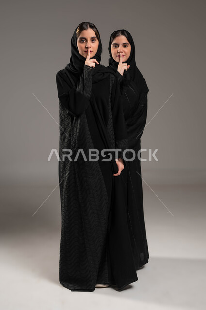 طلب السكوت والصمت, التوقف عن الكلام, بورتريه لبنتان توءم عربيتان سعوديتان خليجيتان ترتديان الحجاب والعباءة, الاشارة بالاصبع الى خفض الصوت والهمس, صورة الجسم بالطول الكامل, خلفية رمادية