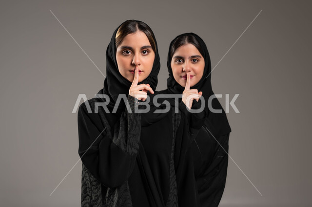 التوقف عن الكلام, طلب السكوت والصمت, بورتريه مقرب لبنتان توءم عربيتان سعوديتان خليجيتان ترتديان الحجاب والعباءة, الاشارة بالاصبع الى خفض الصوت والهمس, خلفية رمادية