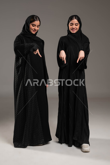 حركات تدل على المكان، الإشارة إلى شيء ما بأصبع السبابة، مفهوم لغة الجسد والحركات والإشارات، الإشارة بالأصابع للأسفل الى شيء ما بايماءات السعادة، ، بورتريه لبنتان توءم عربيتان خليجيتان سعوديتان محجبتان ترتديان عباءة سوداء، صورة الجسم بالطول الكامل، خلفية رمادية