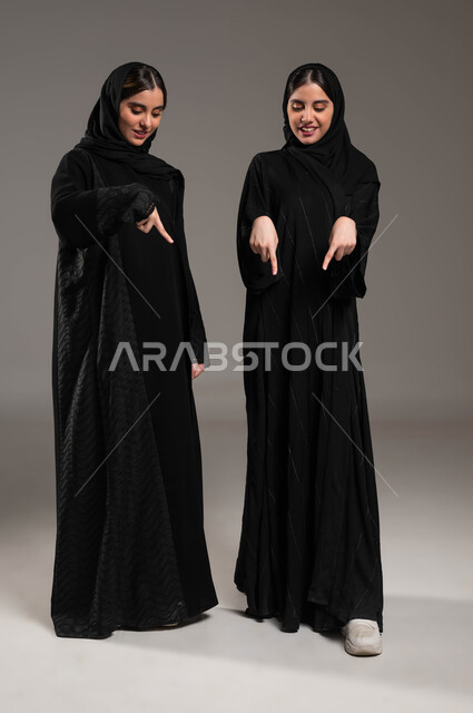 الإشارة بالأصابع للأسفل الى شيء ما بايماءات السعادة، حركات تدل على المكان، الإشارة إلى شيء ما بأصبع السبابة، مفهوم لغة الجسد والحركات والإشارات، بورتريه لبنتان توءم عربيتان خليجيتان سعوديتان محجبتان ترتديان عباءة سوداء، صورة الجسم بالطول الكامل،  خلفية رمادية