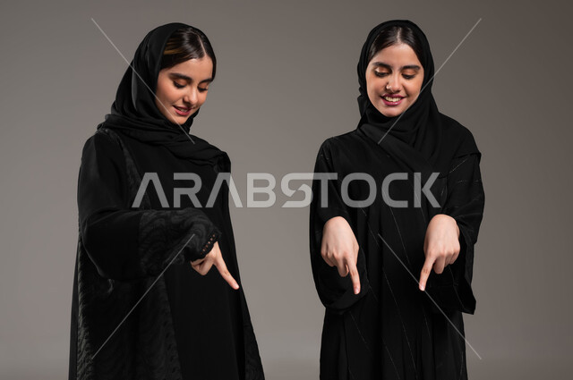 حركات تدل على المكان، الإشارة إلى شيء ما بأصبع السبابة، مفهوم لغة الجسد والحركات والإشارات، بورتريه مقرب لبنتان توءم عربيتان خليجيتان سعوديتان محجبتان ترتديان عباءة سوداء، الإشارة بيديهم للأسفل الى شيء ما بايماءات السعادة , ، خلفية رمادية