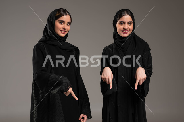 النظر إلى الكاميرا بايماءات السعادة، بورتريه مقرب لتوأم بنات عربيات خليجيات سعوديات ترتديان العباءة والحجاب، حركات تدل على المكان، الإشارة إلى شيء ما في الأسفل بأصبع السبابة، مفهوم لغة الجسد والحركات والإشارات، خلفية رمادية