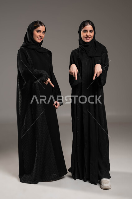 تعابير السعادة في الإشارة إلى شيء ما في الأسفل بأصبع السبابة، النظر إلى الكاميرا بايماءات السعادة، بورتريه لتوأم بنات عربيات خليجيات سعوديات ترتديان العباءة والحجاب، حركات تدل على المكان، مفهوم لغة الجسد والحركات والإشارات، صورة الجسم بالطول الكامل، خلفية رمادية