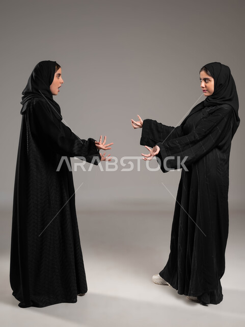 مفهوم النقاش الحاد والمواجهة بين الأخوات، تعابير التعصب والتوتر الانفعالي، بورتريه لتوأم بنات عربيات خليجيات سعوديات محجبات تقفان مقابل بعضهما وتتجادلان بإيماءات الغضب وإلقاء اللوم، صورة الجسم بالطول الكامل، خلفية رمادية