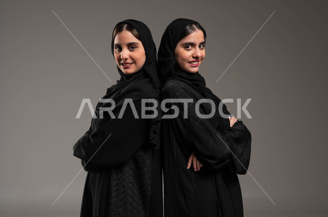 الوقوف باستقامة، الثقة بالنفس والشعور بالقوة, بورتريه مقرب لأختان توءم عربيتان خليجيتان سعوديتان محجبتان ترتديان عباءة سوداء, تقفان بأيدي متقاطعة, ايماءات الفرح والسرور,  خلفية رمادية