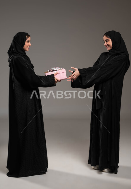 إيماءات الخجل و السرور لتلقي هدية، التعبير عن المحبة والألفة بين الأخوات, تقديم الهدايا والتهنئات، أعياد ومناسبات ومفاجآت سعيدة, بورتريه لشابة عربية خليجية سعودية محجبة ترتدي عباءة سوداء تحمل بيدها علبة هدايا تقوم بتقديمها لأختها، صورة لكامل الجسم، خلفية رمادية