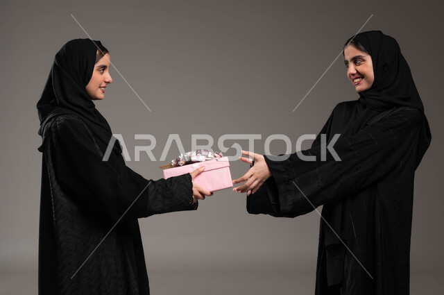 إيماءات السرور لتقديم الهدايا التذكارية، التعبيرعن المحبة والألفة بين الأخوات, أعياد ومناسبات ومفاجآت سعيدة, بورتريه مقرب لشابة عربية خليجية سعودية محجبة ترتدي عباءة سوداء تحمل بيدها علبة هدايا تقوم بتقديمها لأختها، خلفية رمادية