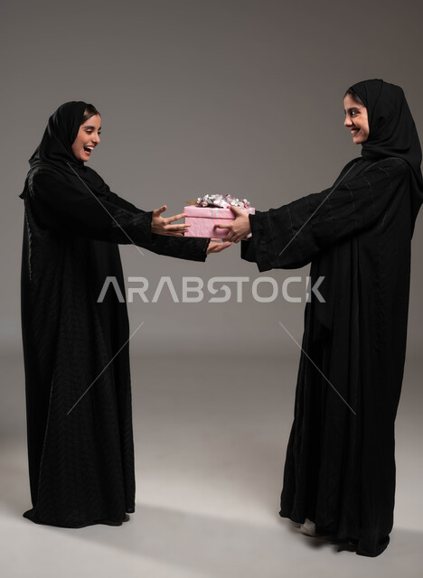 إيماءات السرور و السعادة لتلقي هدية،التعبير عن المحبة والألفة بين الأخوات, تقديم الهدايا والتهنئات، أعياد ومناسبات ومفاجآت سعيدة, بورتريه لشابة عربية خليجية سعودية محجبة ترتدي عباءة سوداء تحمل بيدها علبة هدايا تقوم بتقديمها لأختها، صورة لكامل الجسم، خلفية رمادية