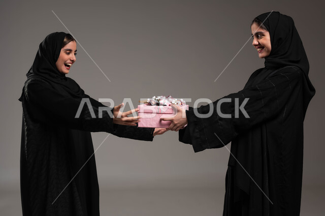 أعياد و مناسبات و مفاجآت سعيدة، إيماءات السرور والسعادة لتلقي الهدايا التذكارية، التعبيرعن المحبة والألفة بين الأخوات، بورتريه مقرب لشابة عربية خليجية سعودية محجبة ترتدي عباءة سوداء تحمل بيدها علبة هدايا تقوم بتقديمها لأختها، خلفية رمادية