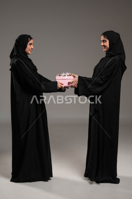التعبير عن المحبة والألفة بين الأخوات, تقديم الهدايا والتهنئات، أعياد ومناسبات ومفاجآت سعيدة, بورتريه لشابتان عربيتان خليجيتان سعوديتان محجبتان ترتديان الثوب عباءة سوداء تحملان علبة هدايا تنظران لبعضهما بإيماءات الخجل والسرور، صورة لكامل الجسم، خلفية رمادية