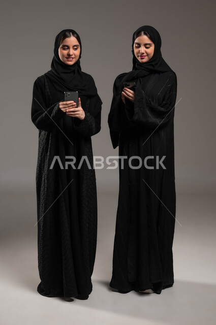 النظر خلسة للهاتف، استخدام الأجهزة والتقنيات الحديثة والمتطورة، التواصل الاجتماعي مع الأهل والاصدقاء عبر الموبايل، بورتريه لشابتان عربيتان خليجيتات سعوديتان محجبتان ترتديان عباءة تحملان بيدهما الجوال، صورة الجسم بالطول الكامل، خلفية رمادية
