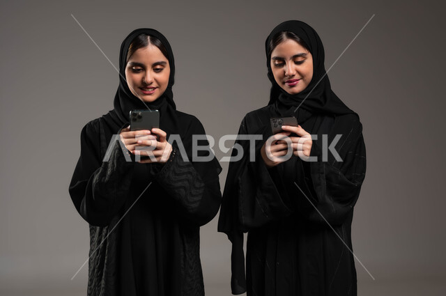 استخدام التقنيات الحديثة، التواصل الاجتماعي و التحدث مع الاصدقاء عبرالموبايل، بورتريه مقرب لشابتان عربيتان خليجيتات سعوديتان محجبتان ترتديان عباءة تحملان بديهما الهاتف تنظران إلى الجوال بتركيز وإندماج، إيماءات السرور و السعادة، خلفية رمادية