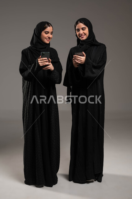 Looking at the mobile phone and sharing happy moments, using modern technologies, social networking and talking with family and friends via the mobile phone, a close-up portrait of two veiled Saudi Gulf Arab women wearing an abaya, holding the phone in their hands, gestures of pleasure and happiness, gray background
