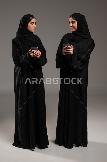 Gestures of teasing, coercion and maliciousness, use of modern and advanced devices and technologies, social communication with family and friends via mobile phone, portrait of two veiled Saudi Arabian Gulf women wearing an abaya, holding a mobile phone in their hand, full-length body photo, gray background